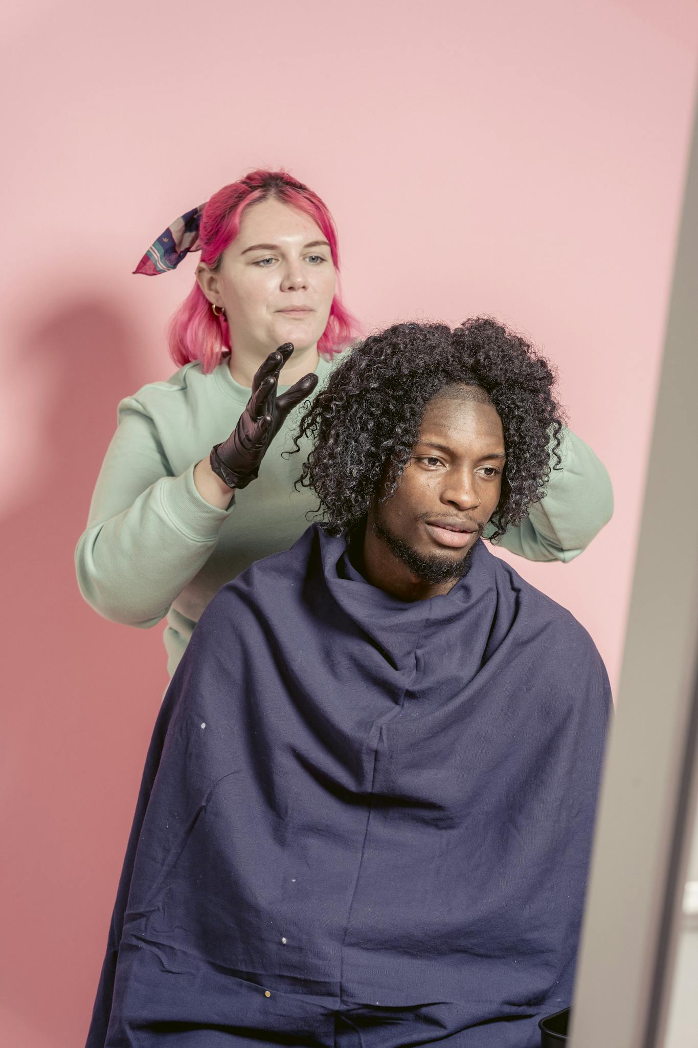 Female hairstylist standing behind bearded African American male client wrapped in cape while touching hair